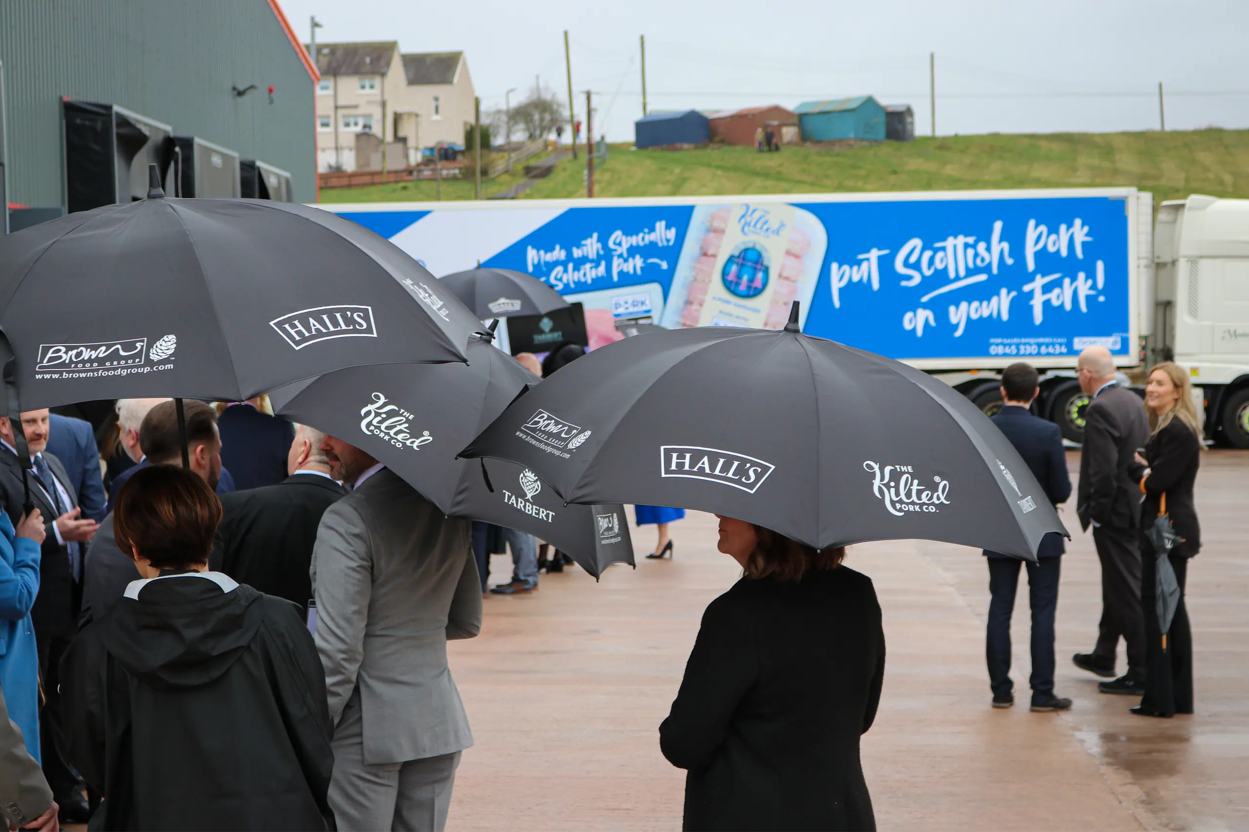 Guests with Hall's and Browns umbrellas gather in the rain outside the Kelloholm factory for the opening of the extension in March 2024